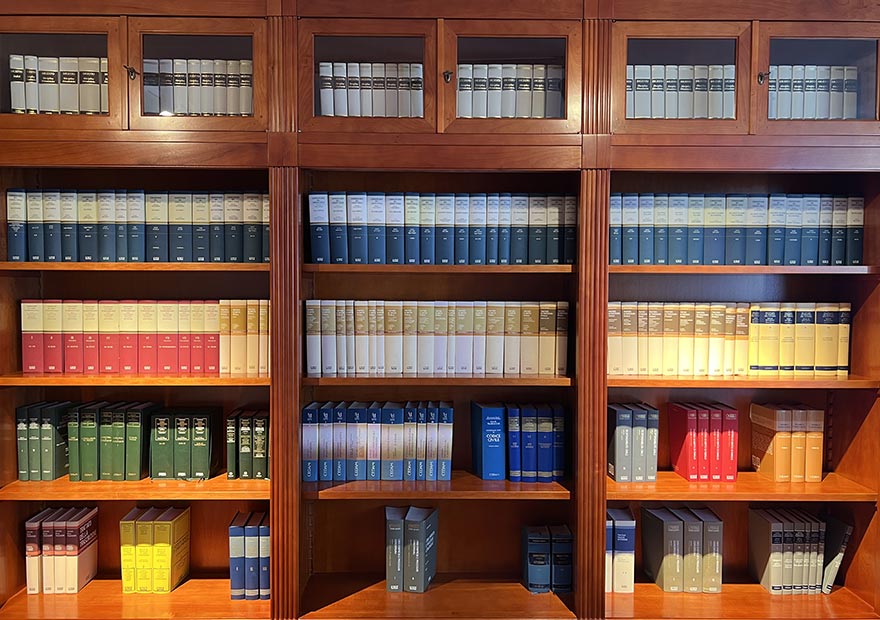 Biblioteca giuridica online - Un portale digitale fluttuante (tipo ologramma) emerge in mezzo a una libreria classica. All’interno del portale si vedono volumi con etichette luminose e accessibili da remoto, mentre una persona li seleziona.